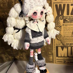 Puede incluir: Una muñeca hecha a mano con un diseño único. La muñeca tiene una cara blanca con mejillas rosadas y ojos grandes. Viste un suéter gris y blanco con la palabra "FASHION", un cuello blanco y zapatos negros. El pelo de la muñeca está peinado en grandes coletas blancas y esponjosas.