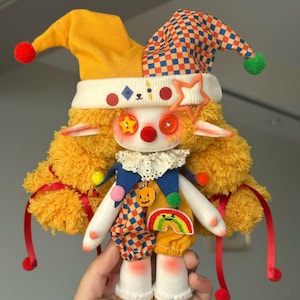 May include: A handmade plush doll dressed as a jester. The doll has a yellow and checkered hat, a white collar, and a colorful outfit with a rainbow and pumpkin patch. The doll's face has button eyes and a red nose.