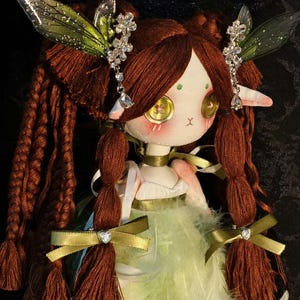 May include: A handmade doll with a whimsical fairy design. The doll has long, braided brown hair, button eyes, and a green and white feathered skirt. It is adorned with decorative crystal accents and fairy wings.