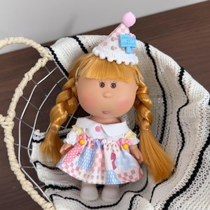Può includere: Una bambola con trecce bionde che indossa un vestito bianco con una gonna patchwork rosa, blu e bianca. La bambola ha un cappello da festa con un pompon rosa e la scritta "notes on cherish". La bambola è in un cestino di filo metallico.