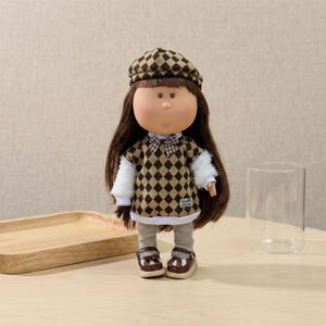 Può includere: Bambola con lunghi capelli castani, indossa un maglione marrone e beige a rombi, un berretto abbinato e scarpe marroni. La bambola ha un papillon e leggings grigi. La bambola è su una superficie chiara.