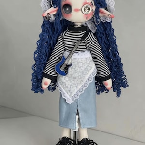 Puede incluir: Una muñeca artesanal con largo cabello azul rizado, tocado de encaje blanco, camisa de manga larga a rayas blancas y negras, delantal de encaje floral blanco y falda azul claro. La muñeca tiene un ojo de botón negro y un monóculo.