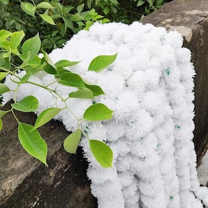 May include: A cascading garland of white, fluffy, pom-pom-like flowers. The garland is draped over a dark, textured surface. Bright green leaves and stems from a nearby plant add a contrasting element to the scene.