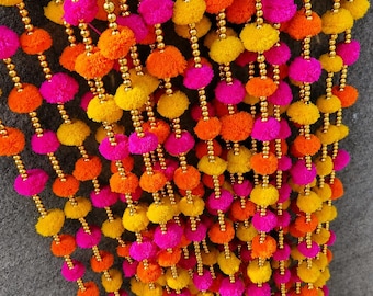 May include: A decorative hanging garland featuring multiple vertical strands of vibrant yellow, orange, and hot pink pom-poms interspersed with small, shiny gold-toned beads. The bottom of each strand is finished with a small, metallic gold bell.