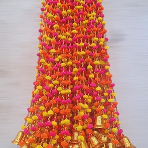 May include: A decorative garland with strands of pink, yellow, and orange pom-poms. The garland is decorated with gold-coloured bells at the bottom, creating a festive and vibrant appearance. Suitable for festive decorations.