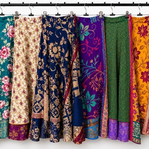 May include: A row of six long skirts hanging from a black metal rack with silver clips. The skirts feature various patterns including floral prints, geometric designs, and intricate borders in teal, cream, navy blue, purple, green, and yellow.
