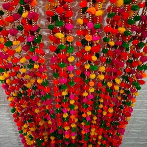 Peut inclure: Une guirlande de pompons multicolores. Elle présente des pompons rouges, roses, jaunes, verts et oranges. Les pompons sont enfilés avec des perles dorées, créant un élément décoratif festif et joyeux, parfait pour les célébrations.