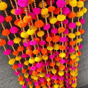 Puede incluir: Una guirnalda decorativa colgante que presenta múltiples hilos verticales de pompones de lana en colores rosa vibrante, naranja y amarillo, intercalados con pequeñas cuentas doradas brillantes y campanas de metal dorado en la base.