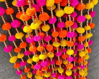 May include: A decorative hanging garland featuring multiple vertical strands of vibrant pink, orange, and yellow yarn pom-poms interspersed with small, shiny gold-colored beads, finished with metallic gold bells at the bottom of each strand.
