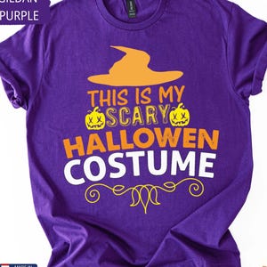 May include: A purple t-shirt with the text "THIS IS MY SCARY HALLOWEEN COSTUME" in orange and white. The design includes an orange witch hat and two jack-o'-lanterns. The shirt is made in the USA.