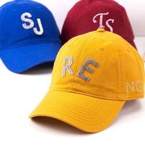 Könnte beinhalten: Eine gelbe Baseballkappe mit den Buchstaben R und E aus silbernen Strasssteinen auf der Vorderseite und N und G an der Seite. Im Hintergrund eine blaue Kappe mit silbernen Strassbuchstaben S und J sowie eine kastanienbraune Kappe mit Buchstaben Ts.