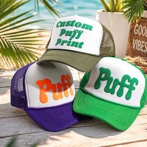 Könnte beinhalten: Drei Trucker-Caps werden gezeigt, jede mit weißer Vorderseite und Mesh-Rückseite. Eine ist olivgrün mit "Custom Puff Print" in Grün. Eine andere ist lila mit "Puff" in Orange. Die dritte ist grün mit "Puff" in Grün.