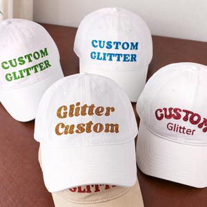 Può includere: Quattro cappellini da baseball bianchi con testo glitterato. Un cappellino ha il testo verde CUSTOM GLITTER, uno il testo blu CUSTOM GLITTER, uno il testo dorato Glitter Custom e uno il testo rosso CUSTOM Glitter.