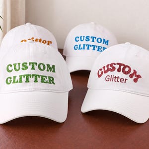 May include: Four white baseball caps featuring the text CUSTOM GLITTER in various colors. One cap has green glitter text, one has red glitter text, one has blue glitter text, and one has orange glitter text. Each cap has a curved brim and a structured crown.
