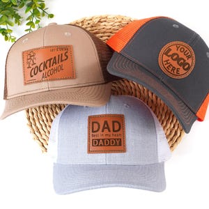 Könnte beinhalten: Drei Trucker-Caps mit Lederaufnähern. Eine beige Kappe hat eine braune Mesh-Rückseite und trägt die Aufschrift "COCKTAILS ALCOHOL". Eine weitere ist grau mit einem orangefarbenen Mesh-Rücken und einem Aufnäher "YOUR LOGO HERE". Die dritte ist grau und weiß mit einem "DAD"-Aufnäher.