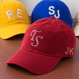 Op de afbeelding: Een donkerrode honkbalpet met de letters TS in doorzichtige strass-steentjes op de voorkant en de letters JK in strass-steentjes aan de zijkant. Andere honkbalpetten in geel, blauw en beige met strass-letters zijn zichtbaar op de achtergrond.