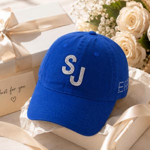 May include: A royal blue baseball cap featuring the letters SJ in clear rhinestones on the front panel. The side of the cap displays additional rhinestone detailing. The cap has a structured crown, a curved brim, and a button on the top centre.