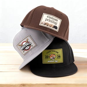Könnte beinhalten: Drei Baseballkappen auf einer Holzoberfläche gestapelt. Die obere Kappe ist braun mit einem Aufnäher mit der Aufschrift "custom printing" und Welpen. Die mittlere Kappe ist grau mit einem Baseball-Club-Logo. Die untere Kappe ist schwarz mit einem Tiger-Logo und dem Text "Your Tagline".