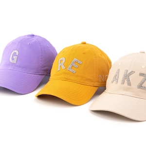 Könnte beinhalten: Drei Baseballkappen in Lavendel, Senfgelb und Beige. Jede Kappe ist auf den Vorderteilen mit strassbesetzten Buchstaben versehen. Die lavendelfarbene Kappe zeigt den Buchstaben G, die gelbe RE und die beige Kappe AKZ.