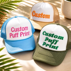Op de afbeelding: Drie truckerpetten met witte voorpanelen en mesh-rug. Eén is blauw met roze tekst "Custom Puff Print", een andere is bruin met oranje tekst "Custom", en de derde is groen met groene tekst "Custom Puff Print".