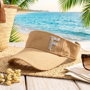 Könnte beinhalten: Beigefarbener Visor mit einem funkelnden Buchstaben "F" aus Strasssteinen. Der Visor liegt auf einer gewebten Matte, mit einer Strandkulisse im Hintergrund, einschließlich Palmen und Meer.