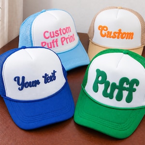 Könnte beinhalten: Vier Trucker-Caps mit weißen Schaumstoff-Frontpaneelen und Netzrücken. Die blaue Kappe zeigt den dunkelblauen Schriftzug Your text. Die hellblaue hat den rosa Schriftzug Custom Puff Print. Die beige hat den orangen Schriftzug Custom. Die grüne hat den grünen Schriftzug Puff.