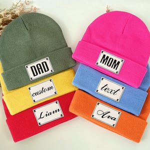May include: A collection of colorful beanies in green, yellow, red, pink, blue, and orange. Each beanie features a rectangular patch with text, including "DAD", "MOM", "custom", "text", "Liam", and "Ava".