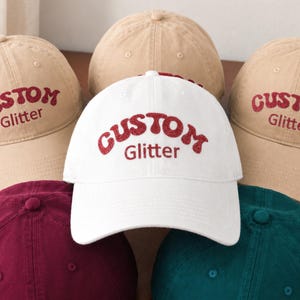 May include: A collection of five baseball caps in white, beige, maroon, and teal. Each cap features the text CUSTOM Glitter embroidered in red glittery lettering on the front panel. The caps have a structured crown, a curved brim, and a button top.