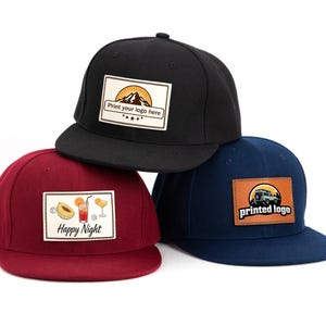 Könnte beinhalten: Drei Baseballkappen in Schwarz, Burgunderrot und Marineblau. Jede Kappe hat einen rechteckigen Aufnäher mit einem anderen Design. Auf der schwarzen Kappe steht "Print your logo here". Auf der bordeauxroten Kappe steht "Happy Night". Auf der marineblauen Kappe steht "printed logo".
