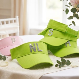 Könnte beinhalten: Eine Auswahl an Sonnenblenden in Rosa und Grün, jeweils mit funkelnden Initialen versehen. Die Sonnenblenden sind auf einem Tisch angeordnet, was auf ein Thema der Personalisierung hindeutet. Die hellen Farben und Verzierungen verleihen den Sonnenblenden ein stilvolles Aussehen.