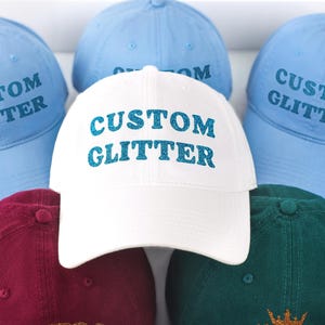 May include: A collection of baseball caps in various colors including white, light blue, maroon, and dark green. The white cap features the text CUSTOM GLITTER in blue glitter, while the light blue caps also display the same text.