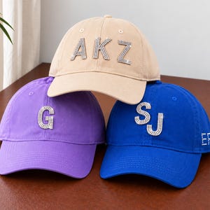 May include: Three baseball caps in beige, purple, and blue. The beige cap features rhinestone letters AKZ. The purple cap displays a single rhinestone letter G. The blue cap shows rhinestone letters SJ on the front and EP on the side panel.