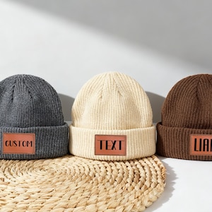 Könnte beinhalten: Drei Strickmützen in Grau, Creme und Braun, jede mit einem rechteckigen Lederaufnäher. Die graue Mütze hat "CUSTOM" auf dem Aufnäher, die cremefarbene "TEXT" und die braune "LIAM". Die Mützen liegen auf einer gewebten Matte.