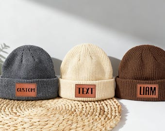 May include: Three knit beanies in gray, cream, and brown, each with a rectangular leather patch. The gray beanie has "CUSTOM" on the patch, the cream one says "TEXT", and the brown one says "LIAM". The beanies are on a woven mat.