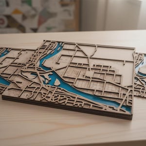 May include: Wooden laser-cut city map art pieces with a river of blue resin. The maps are layered, creating a 3D effect. The pieces are displayed on a light-colored wooden surface.