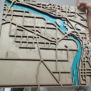May include: A layered wooden map with a light brown finish, depicting a city layout. A blue river runs through the center, with detailed streets and building outlines. The map is held up by a person.