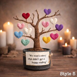 May include: A decorative tree sculpture with a brown trunk and branches, adorned with colorful crochet hearts. The tree sits in a gray pot with a tag that reads, "We were a story that didn't get a happy ending." Candles provide a warm background.