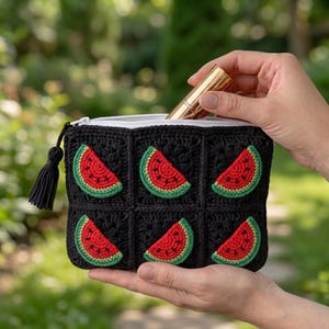 Puede incluir: Un estuche rectangular de ganchillo con fondo negro y seis motivos cuadrados individuales, cada uno con un patrón de rodaja de sandía roja y verde. El estuche incluye una cremallera blanca en la parte superior y un tirador de borla negra.