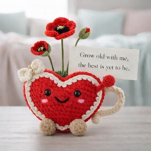 Puede incluir: Una taza en forma de corazón tejida a ganchillo, con una cara sonriente, cuerpo rojo y ribete blanco. Contiene flores de amapola rojas y una tarjeta con el texto: "Envejece conmigo, lo mejor está por venir".