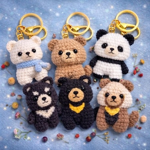 Peut inclure: Collection de six porte-clés ours crochetés de différentes couleurs, dont blanc, marron, noir et un motif panda. Chaque ours a un porte-clés doré. Certains ours ont des détails supplémentaires comme une écharpe bleue ou un bavoir jaune.
