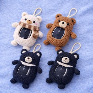 Op de afbeelding: Vier handgemaakte gehaakte beren sleutelhangerhoezen in crème, bruin en marineblauw. De crème beer heeft een bruin-witte sjaal. Elke beer heeft een sleutelopening en een lus. Op een lichtblauwe ondergrond.