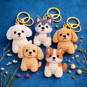 Pode incluir: Cinco porta-chaves de cão em croché com argolas de metal dourado. A coleção inclui um cão branco, um cão tipo husky cinzento e branco, dois cães castanhos claros com orelhas caídas e um cão castanho e branco, todos com olhos e nariz de plástico preto.