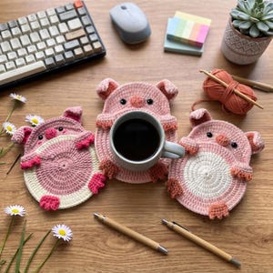 May include: Three round, crocheted pig coasters in shades of light pink and white. Each coaster features small ears, a snout, and tiny feet. One coaster holds a grey ceramic mug filled with black coffee. The coasters are arranged on a wooden surface.