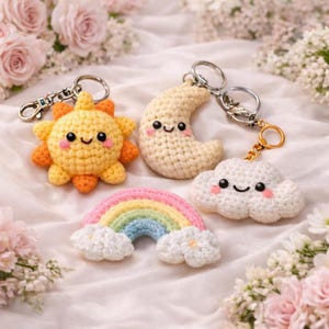 May include: Four crochet keychains: a smiling sun, a crescent moon, a fluffy cloud, and a rainbow with clouds. The sun is yellow and orange, the moon is cream, the cloud is white, and the rainbow has pink, yellow, green, and blue arches. Each keychain has a metal ring.