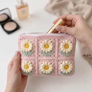 Puede incluir: Un estuche rectangular de ganchillo con fondo rosa claro y seis motivos cuadrados. Cada motivo muestra una margarita tridimensional en relieve con pétalos blancos, un centro amarillo y dos pequeñas hojas de color verde claro en la base. El estuche tiene una cremallera blanca.