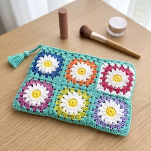 Puede incluir: Un estuche rectangular de ganchillo compuesto por seis cuadrados con motivos de margaritas de centro blanco y amarillo, rodeados de hilo color verde azulado, naranja, magenta, morado y verde lima, con una borla a juego en la esquina.