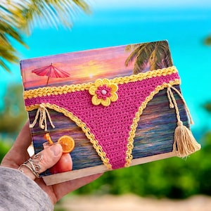 May include: A book cover featuring a tropical beach scene with a sunset, palm trees, and a cocktail. A pink crocheted bikini design with yellow trim and a flower embellishment is on the cover. A tassel and ties are attached to the side.