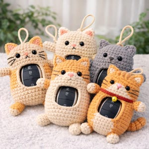 May include: Five crocheted cat-shaped key fob covers in beige, orange, and grey. Each cover has a loop for a keychain and a cutout to display the car key. The cat covers have black embroidered whiskers and pink noses. The key fobs are made of yarn.