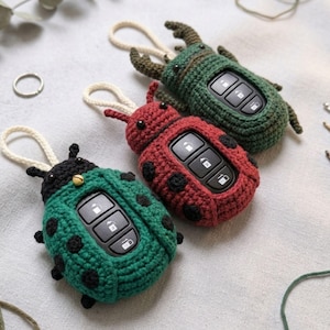 May include: Three handmade crochet key fob covers shaped like insects. One is green with black spots, one is red with black spots, and one is a dark green beetle shape with horns. Each features a white loop and holds a black car key remote.