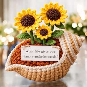 Może przedstawiać: Szydełkowa aranżacja słoneczników w doniczce w kształcie muszli. Słoneczniki mają żółte płatki i brązowe środki, z zielonymi łodygami i liśćmi. Mały znak głosi: „Kiedy życie daje ci cytryny, rób lemoniadę."